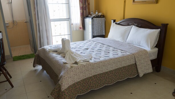 A neatly made bed with a floral bedspread and pillows, a small table with a water bottle, and a window with curtains. - Sabaidee Residence (Bang Bua Thong)