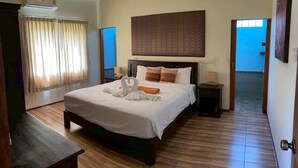 A hotel room with a large bed, a headboard, a bedside table, an air conditioning unit, a curtain, and a view of the outside. - Taratai Villa (Pattaya)