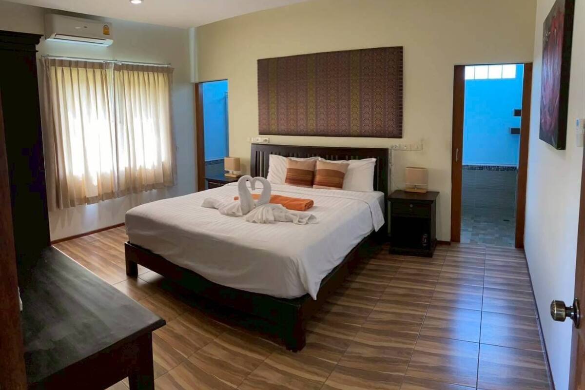 A hotel room with a large bed, a headboard, a bedside table, an air conditioning unit, a curtain, and a view of the outside.