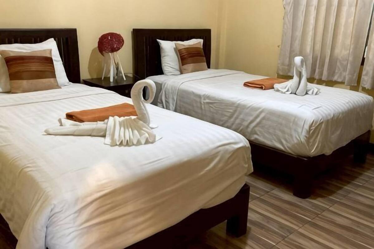 Two double beds with white linens and brown headboards, a small table with a vase, and a window with curtains.