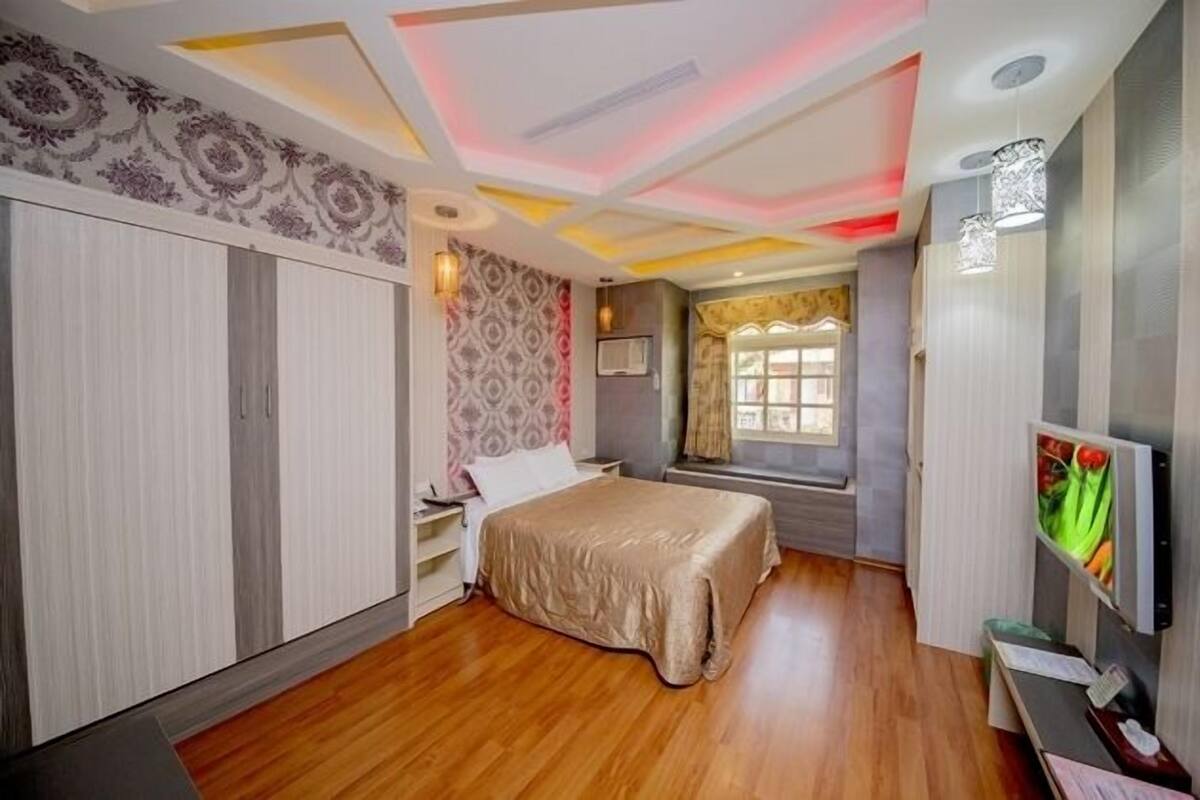 A bedroom with a bed, a desk, a TV, and a window with curtains.