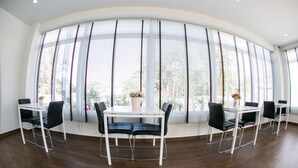 A dining area with a white table and black chairs, large windows with blinds, and a potted plant on the table. - J-House Phetchaburi (Phetchaburi)