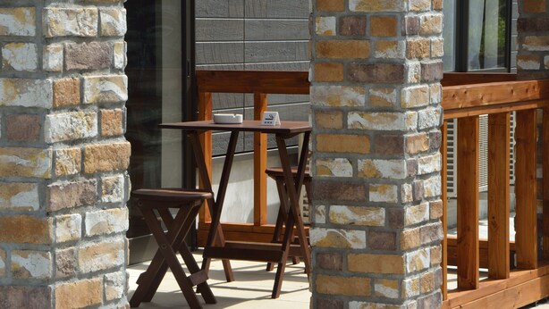 A wooden table and chairs set against a brick wall, with a wooden railing and a window in the background. - Lodge Ocean (Hachijo)