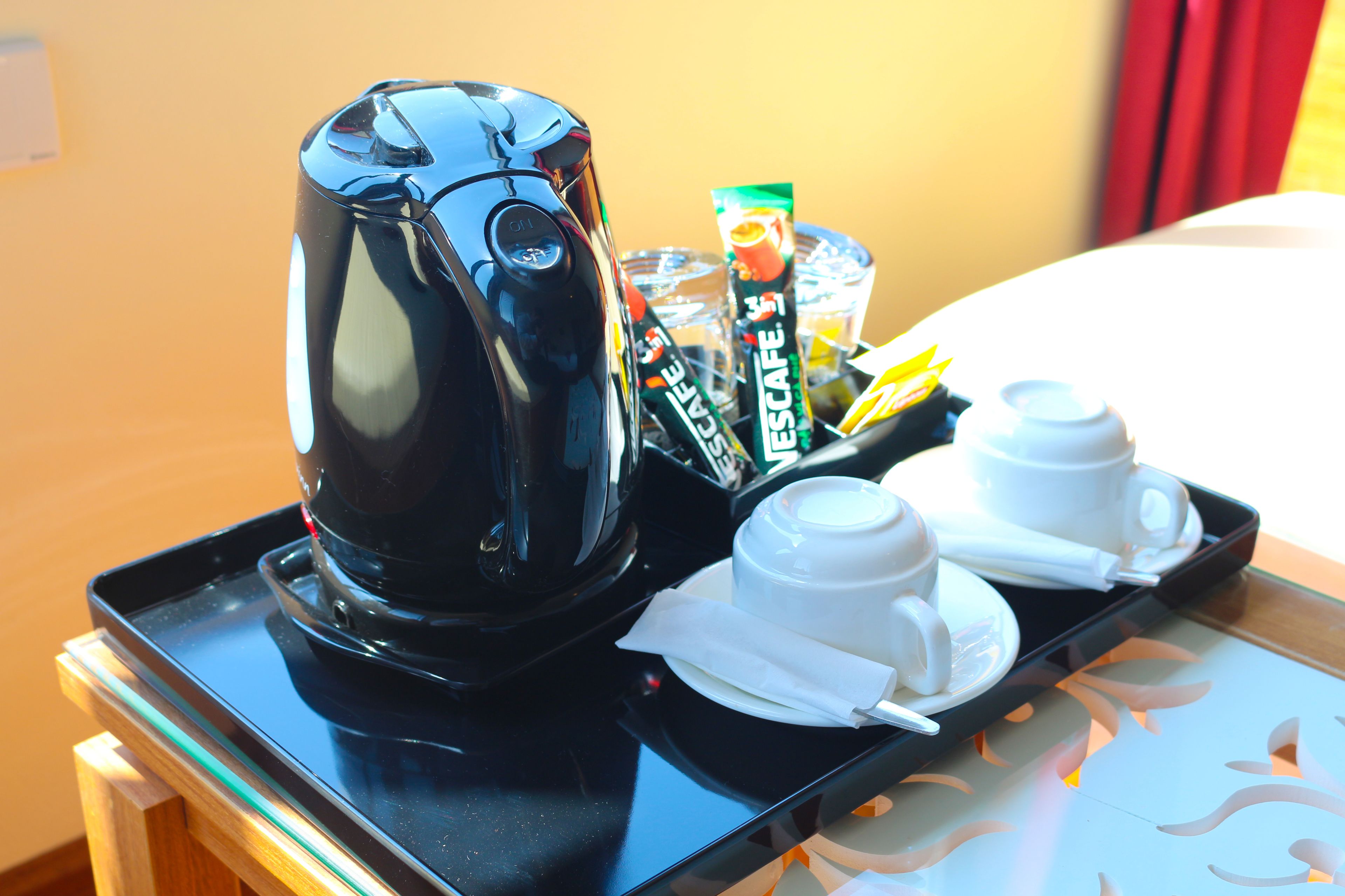 A tray with a black electric kettle, a coffee machine, a cup of coffee, a cup of tea, and a packet of creamer.