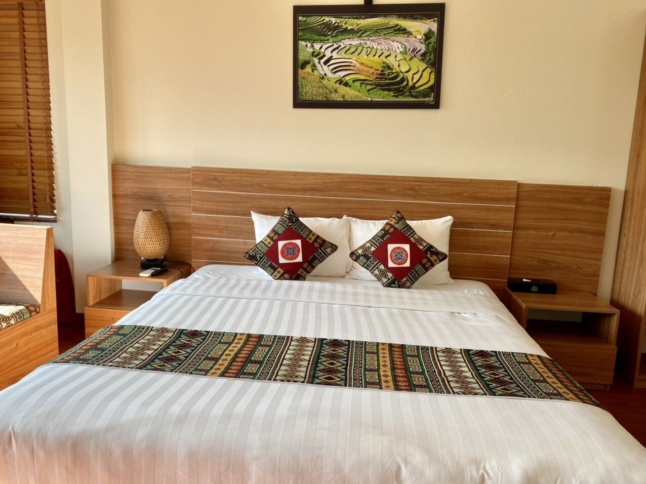 A hotel room with a large bed, wooden headboard, two decorative pillows, a bedside table with a lamp, and a framed picture on the wall.