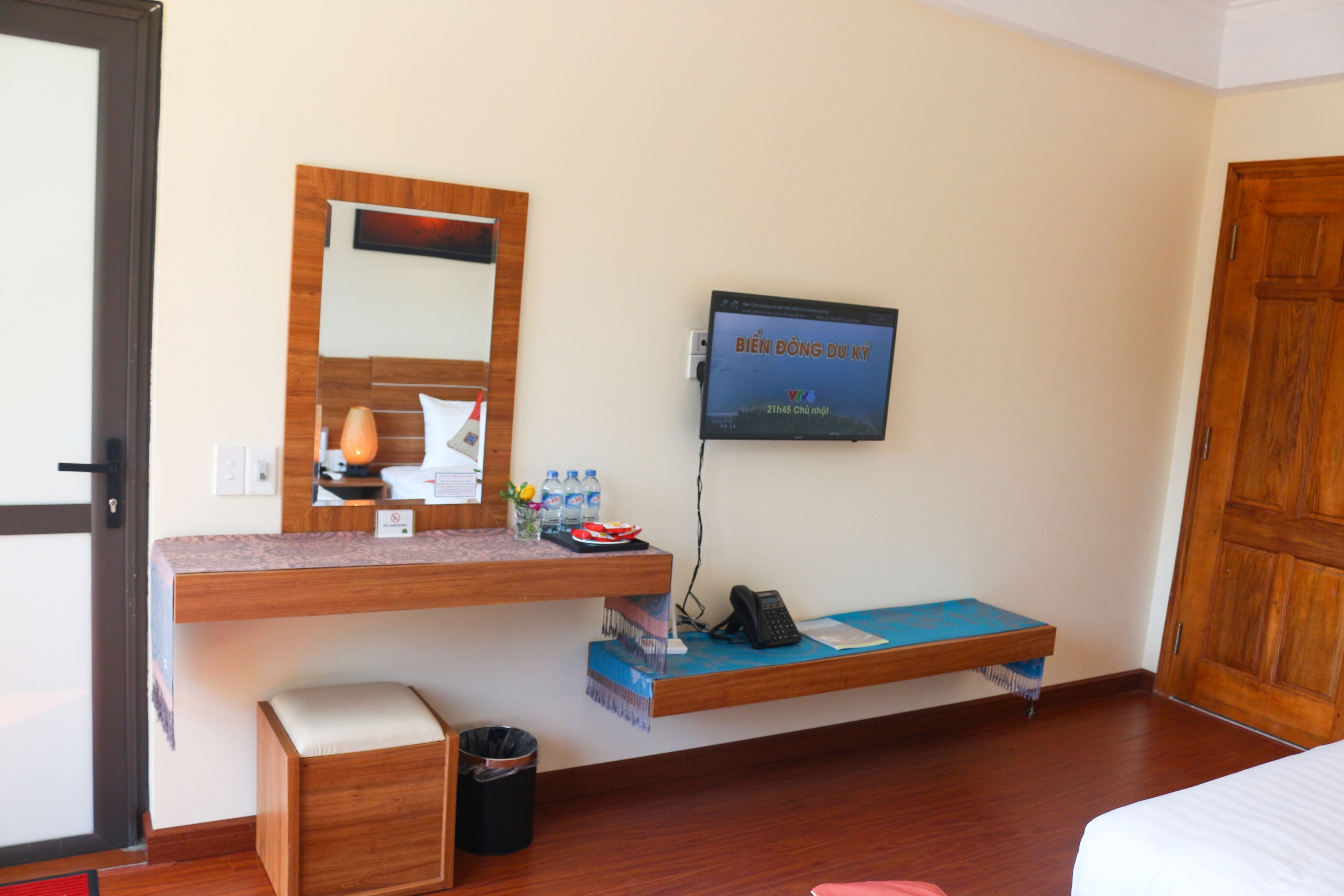 A hotel room with a wooden desk, a television mounted on the wall, a small trash bin, and a wooden door.