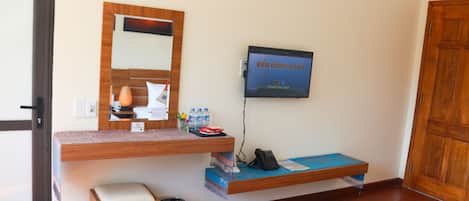 A hotel room with a wooden desk, a television mounted on the wall, a small trash bin, and a wooden door.