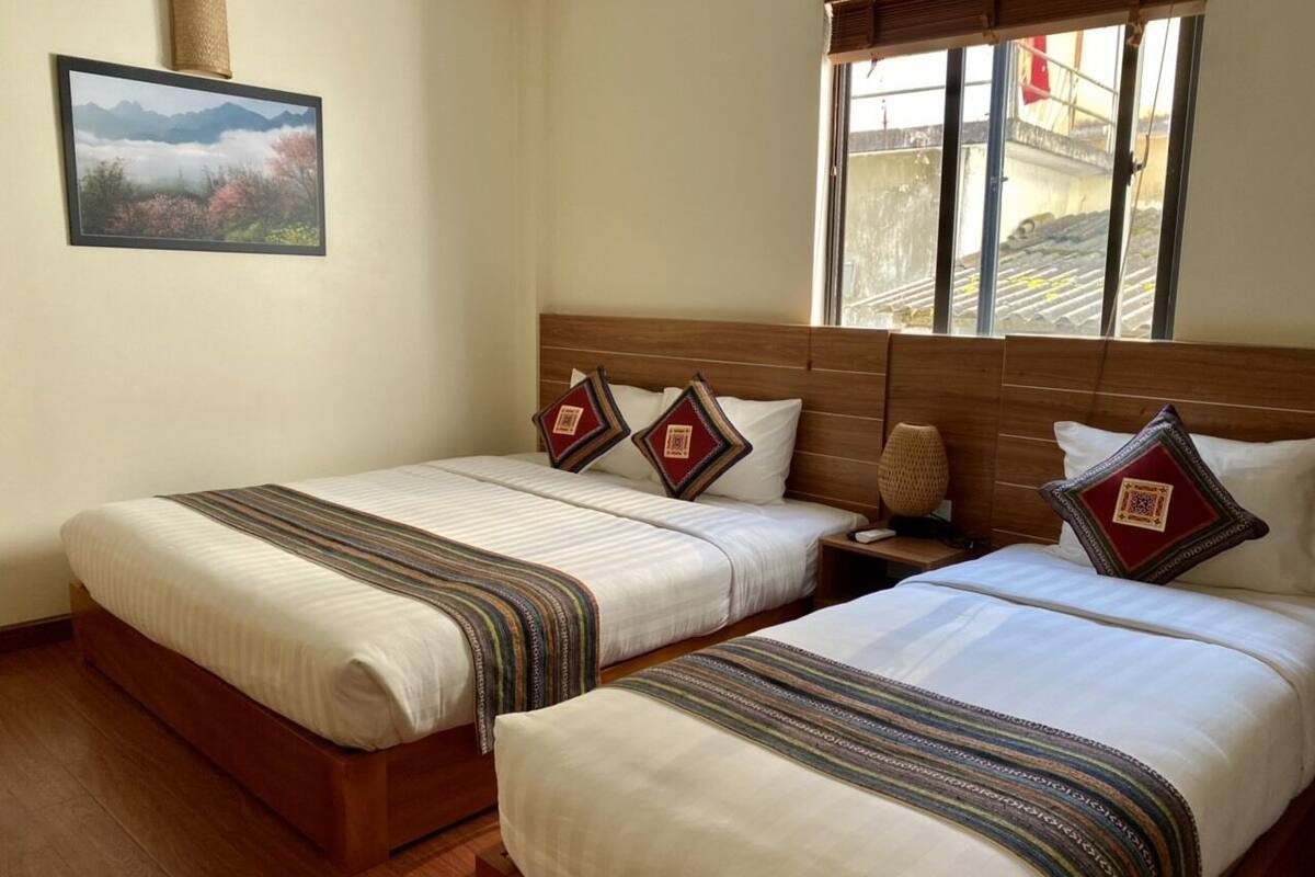 Ett hotellrum med två sängar, var och en med ett randigt överkast, en sänggavel i trä och dekorativa kuddar. En inramad tavla hänger på v äggen, och ett fönster ger utsikt över omgivningen.