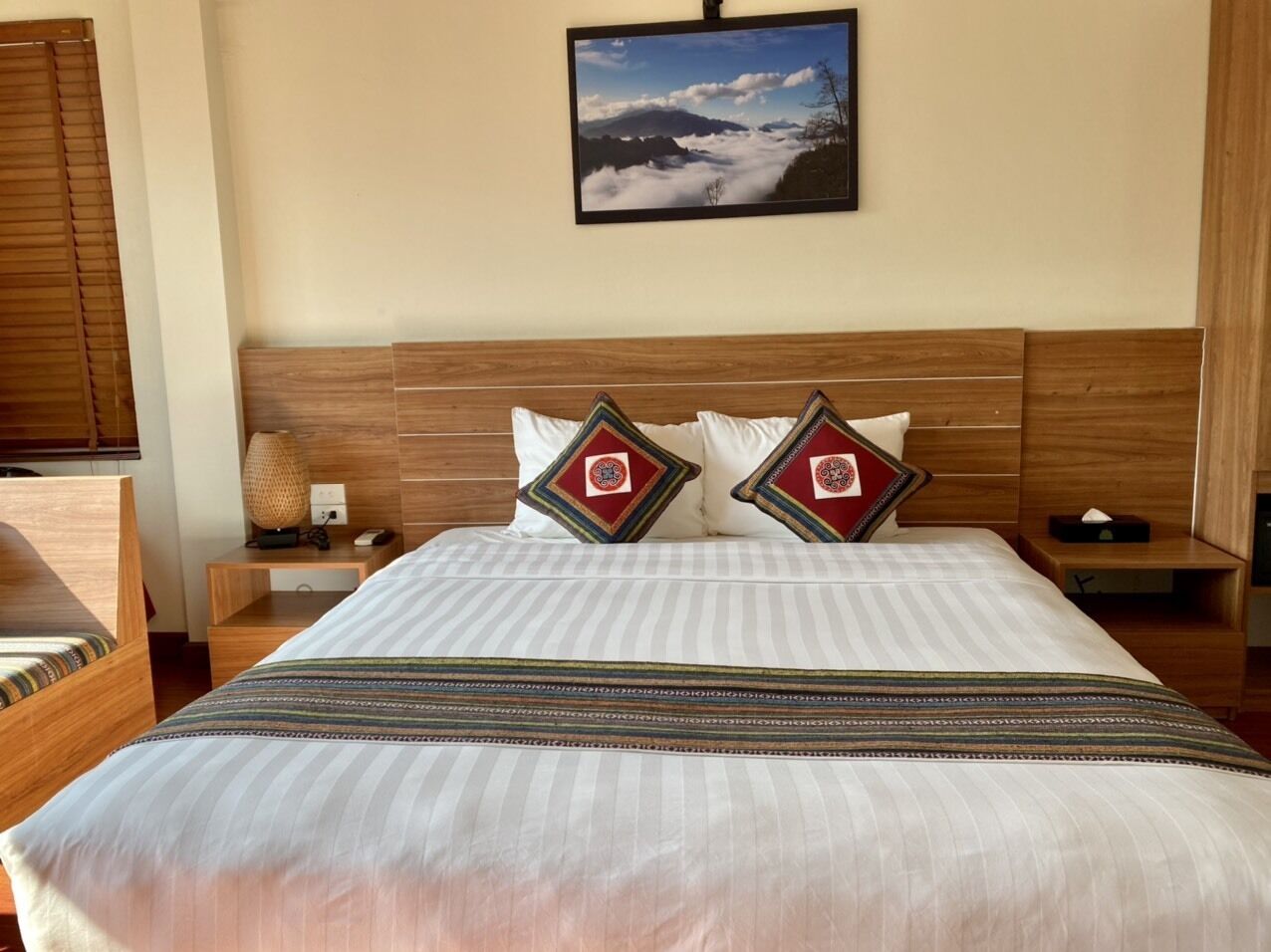 A hotel room with a large bed, two pillows with a red and gold emblem, a nightstand with a lamp, and a framed picture of a mountain landscape on the wall.