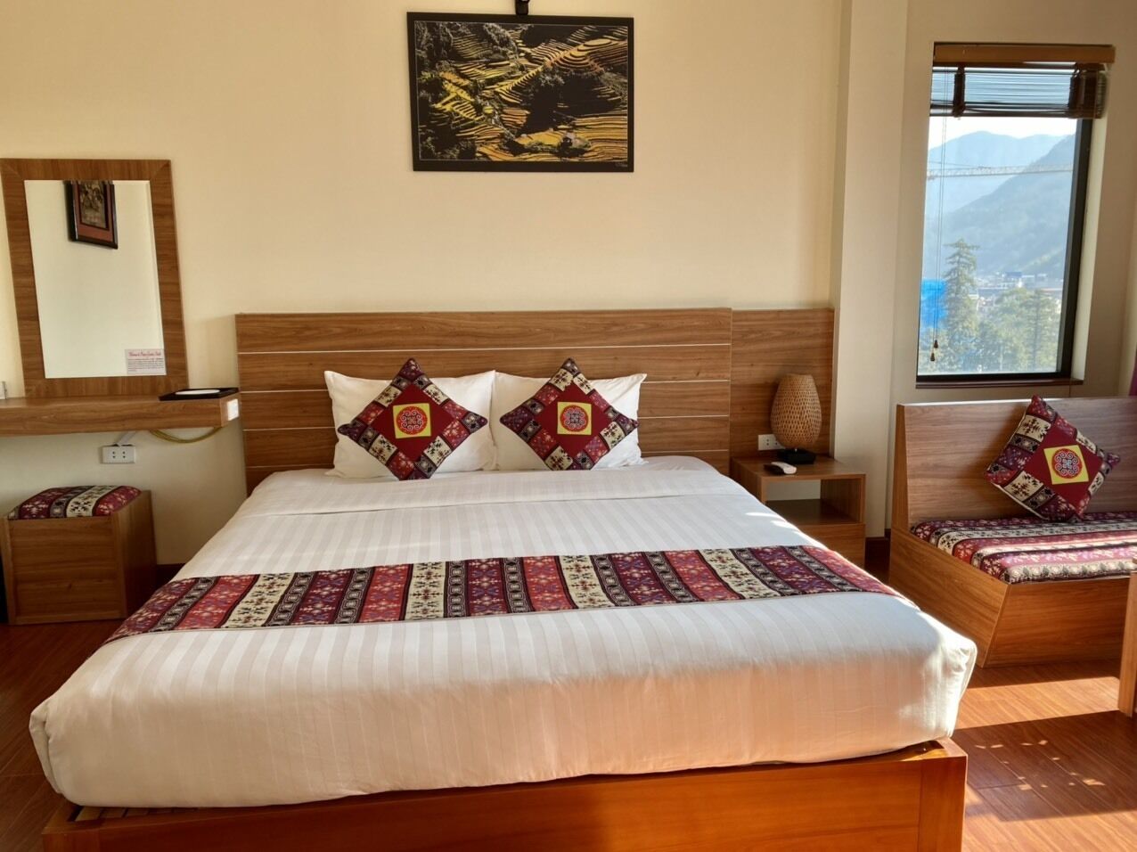 A hotel room with a large bed, a bedside table, a nightstand, a window with a view, and a wall-mounted picture.