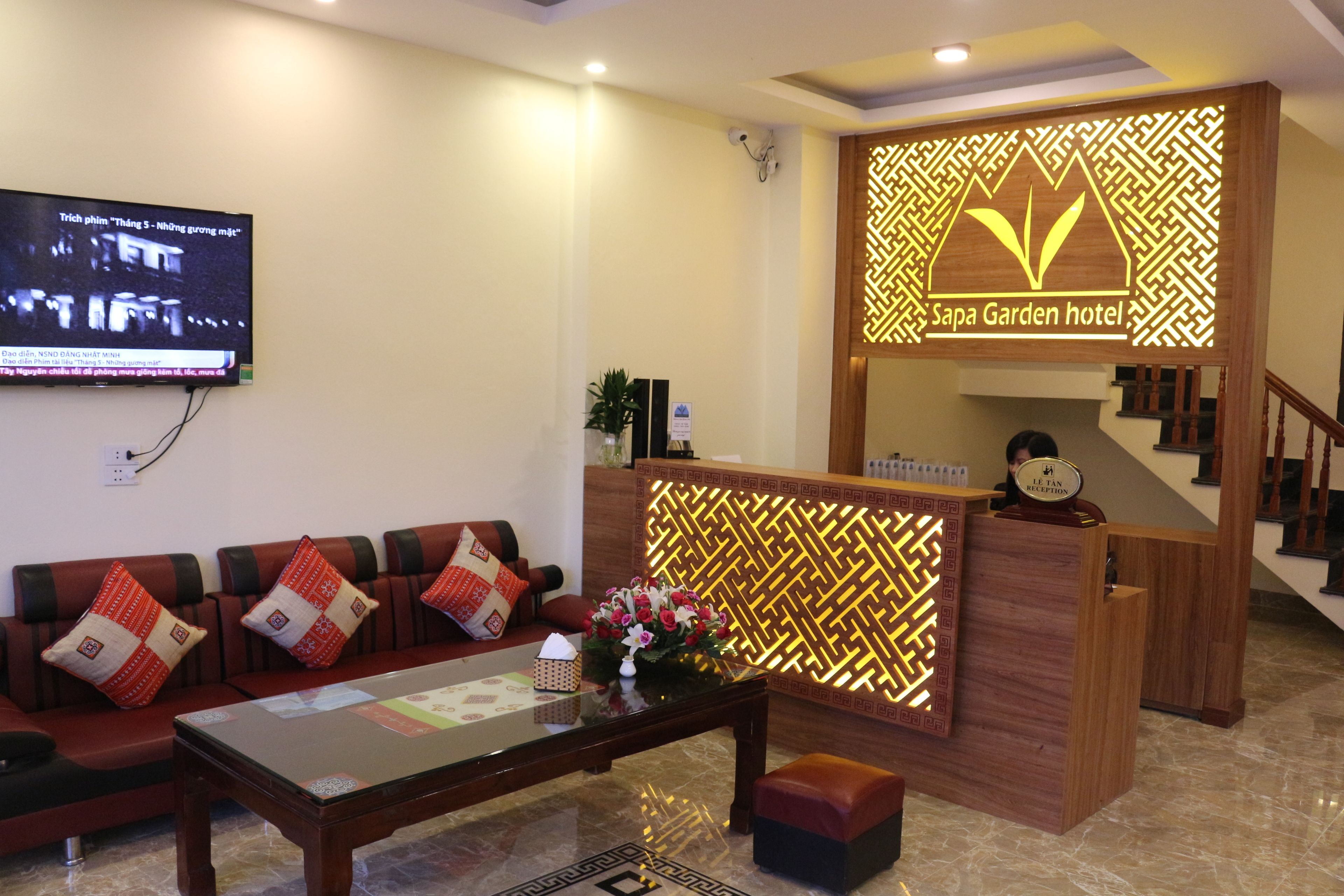 A reception area with a wooden reception desk, a sofa with patterned cushions, a television mounted on the wall, and a decorative wall panel with the hotel's name.
