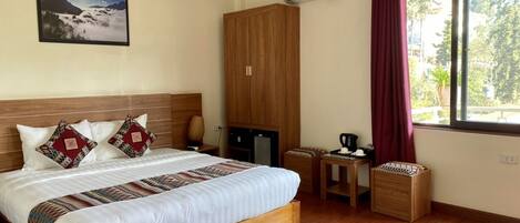 A hotel room with a large bed, a wooden wardrobe, a small desk, a chair, a table, and a window with curtains.