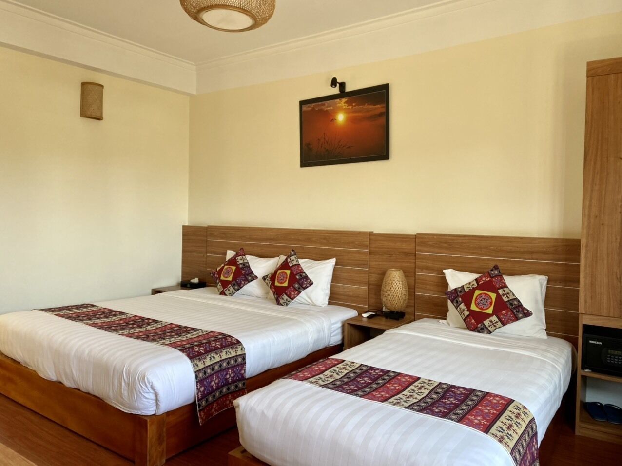 A hotel room with two beds, each with a wooden headboard, white linens, and patterned throw blankets. There is a wall-mounted picture of a sunset, a bedside lamp, and a wooden nightstand with a phone and a small table.