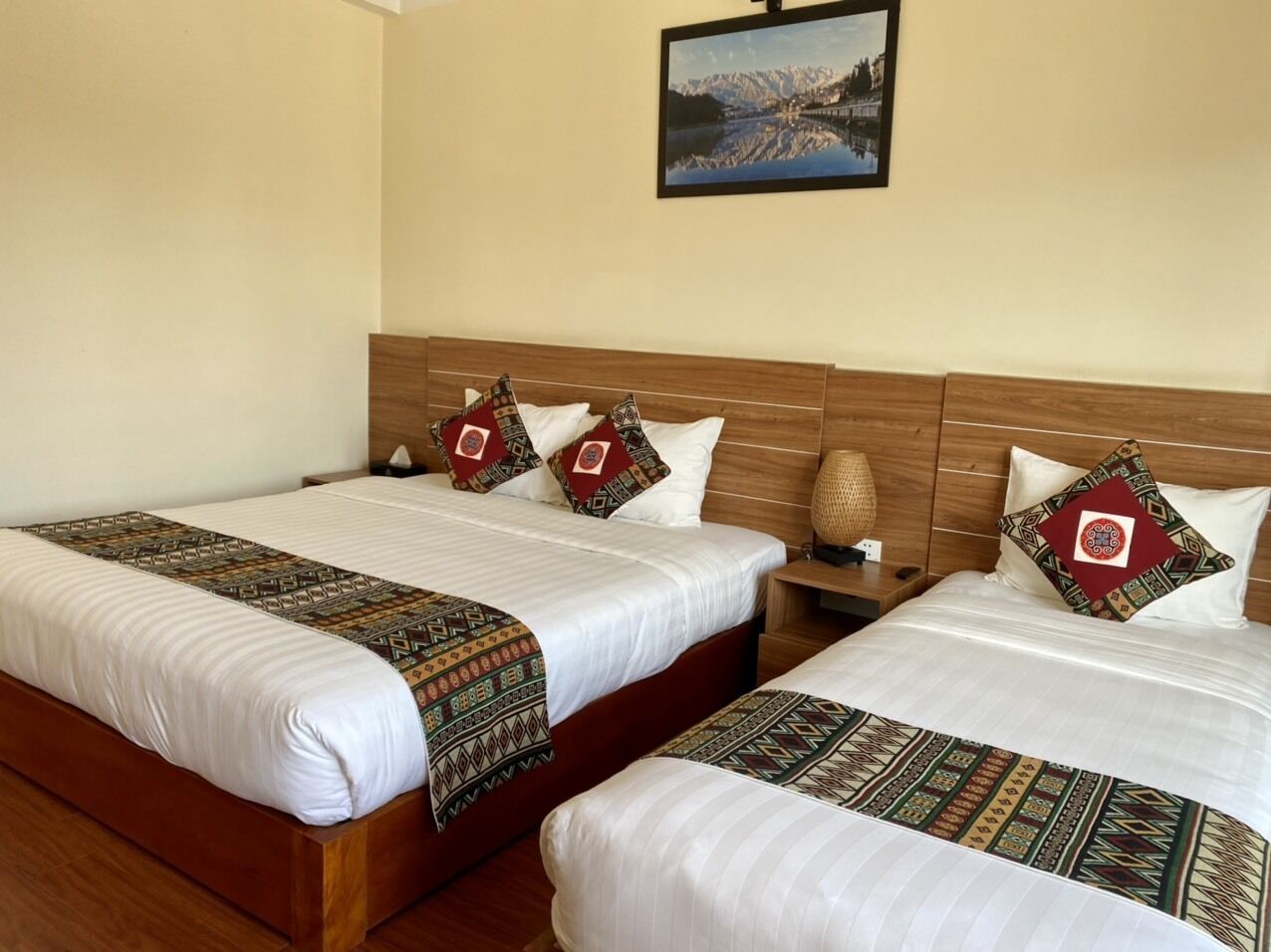 A hotel room with two beds, each with a patterned bedspread and decorative pillows. A framed picture hangs on the wall above the beds.