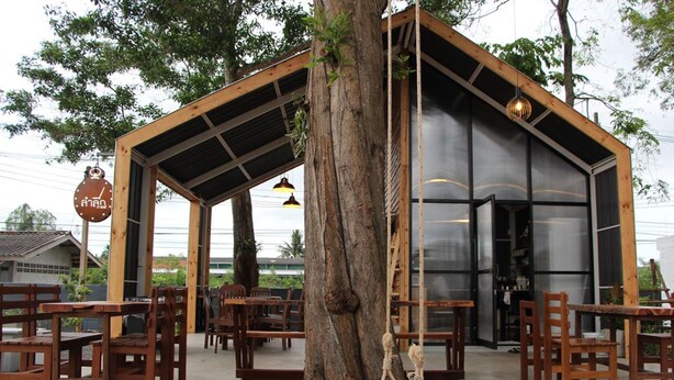 Un espacio al aire libre con mesas y sillas de madera, una hamaca colgada de un árbol, y un edificio moderno con ventanas grandes y techo de metal.