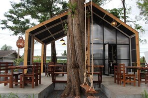 Un espacio al aire libre con mesas y sillas de madera, una hamaca colgada de un árbol, y un edificio moderno con ventanas grandes y techo de metal.