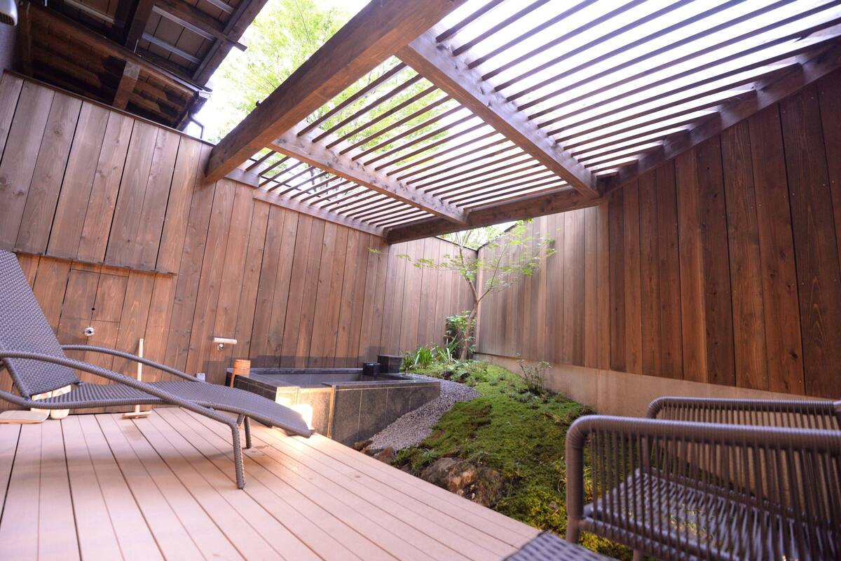 Una terraza de madera con jacuzzi integrado, sillas de descanso y una pérgola.