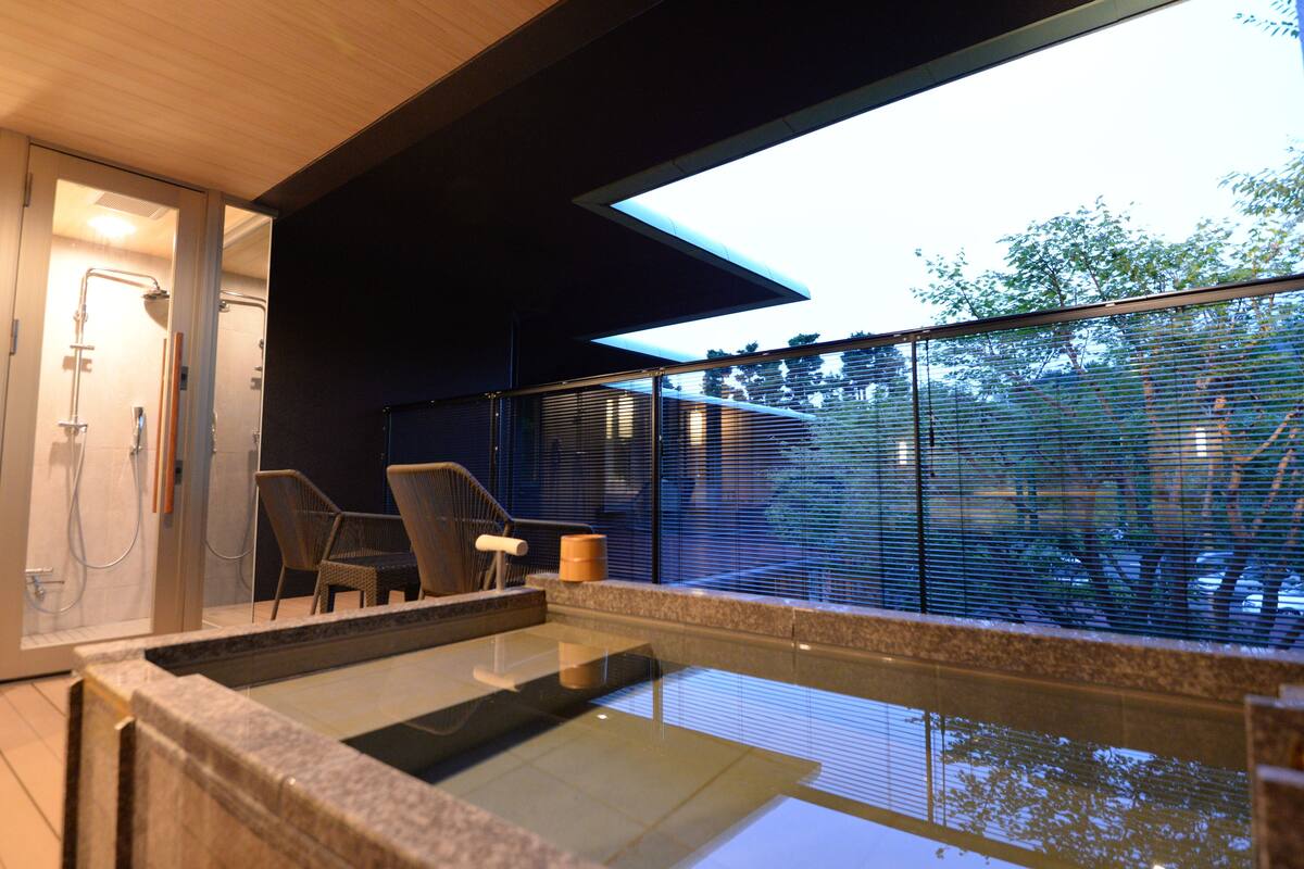 Un baño moderno con ducha envuelta en vidrio, una zona de estar con sillas y vistas amplias por un gran ventanal.