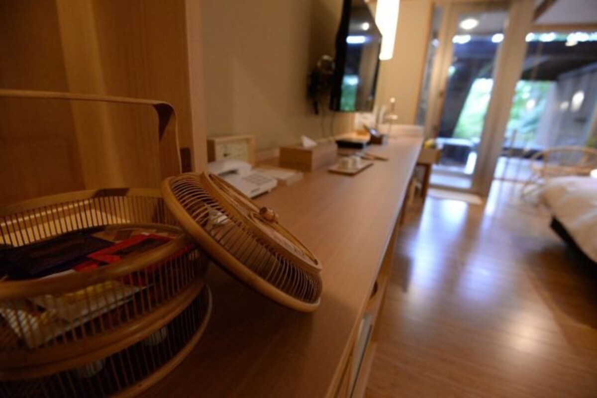 Habitación de hotel con piso de madera, una cama, un escritorio con papeles, un ventilador y una silla.