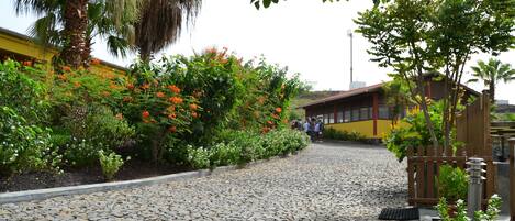 Un camino adoquinado que conduce a un edificio de fachada amarilla, rodeado de vegetación exuberante y plantas florecientes.