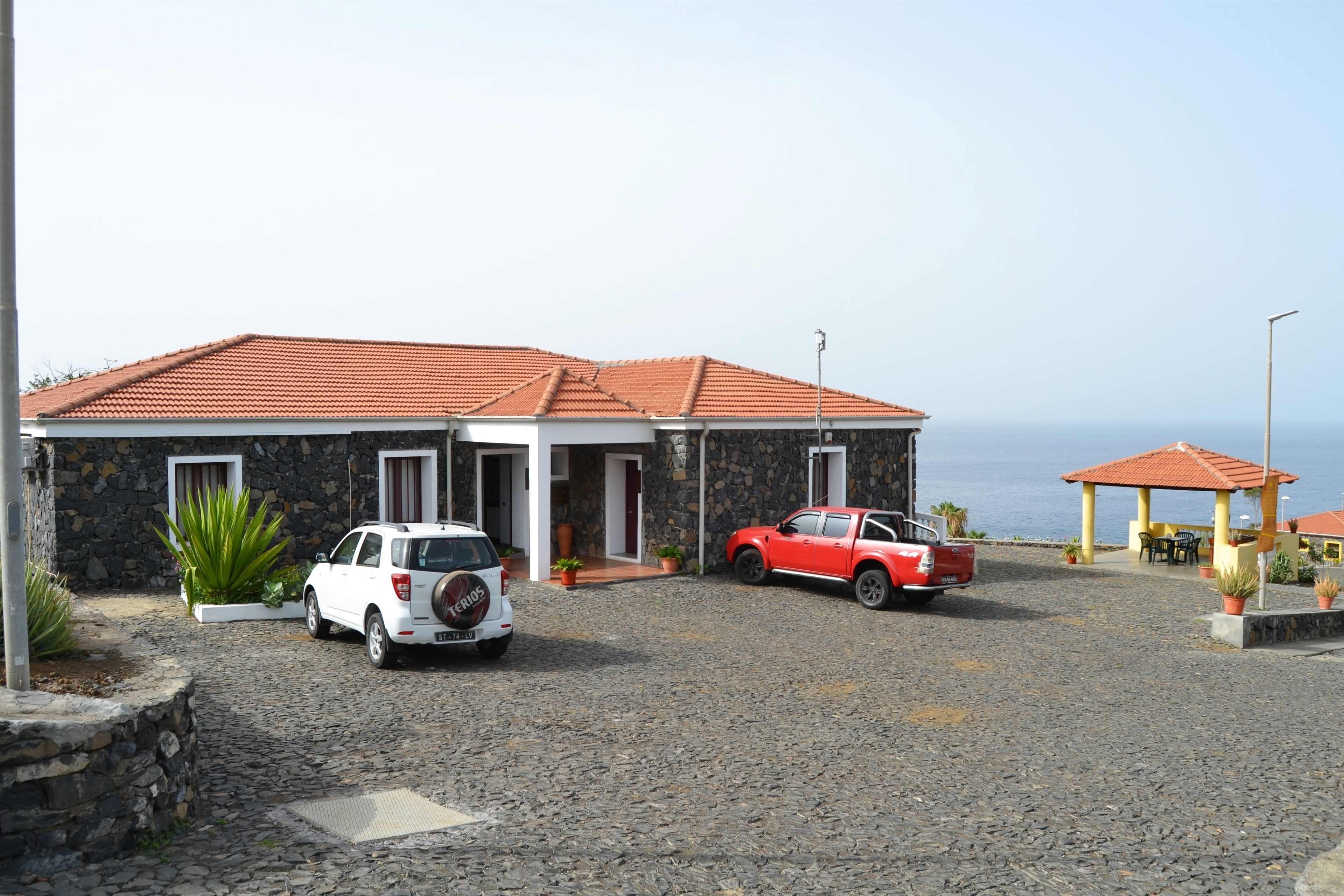 Un edificio con fachada de piedra, dos coches estacionados frente a él y vista al mar.