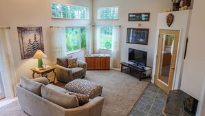 Una sala de estar con un sofá, una mesa de centro, un televisor y una chimenea. Hay ventanas con cortinas, un cuadro en la pared y una puerta.