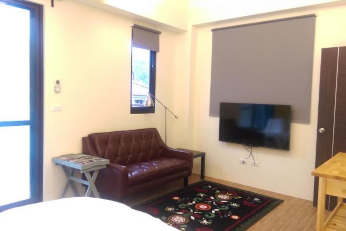 A room with a brown leather sofa, a small side table with magazines, a large flat-screen TV mounted on the wall, and a patterned rug on the floor.