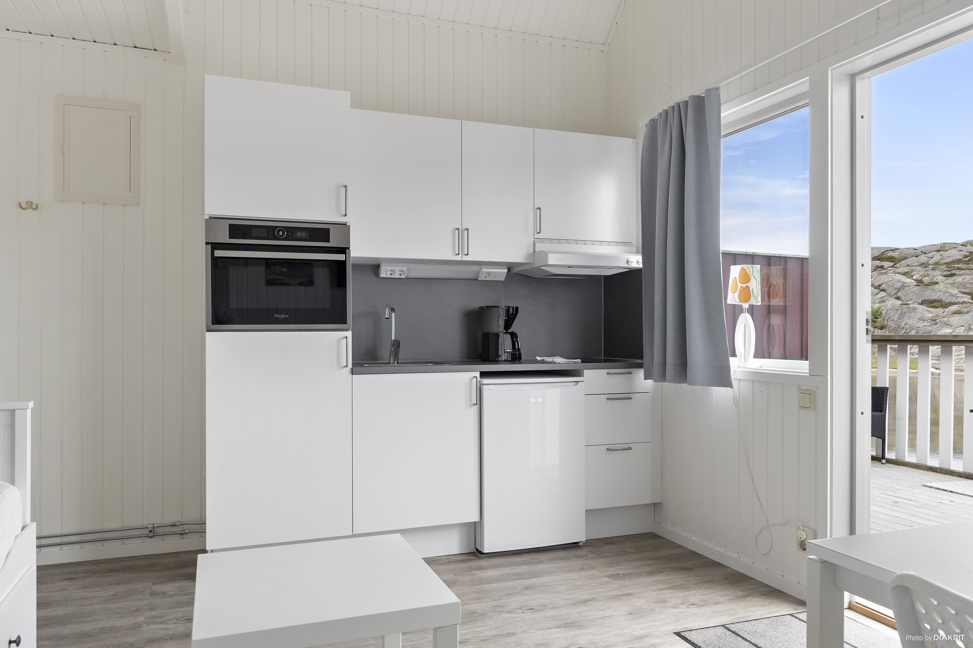 A modern kitchen with white cabinetry, a built-in oven, a dishwasher, and a window with curtains.