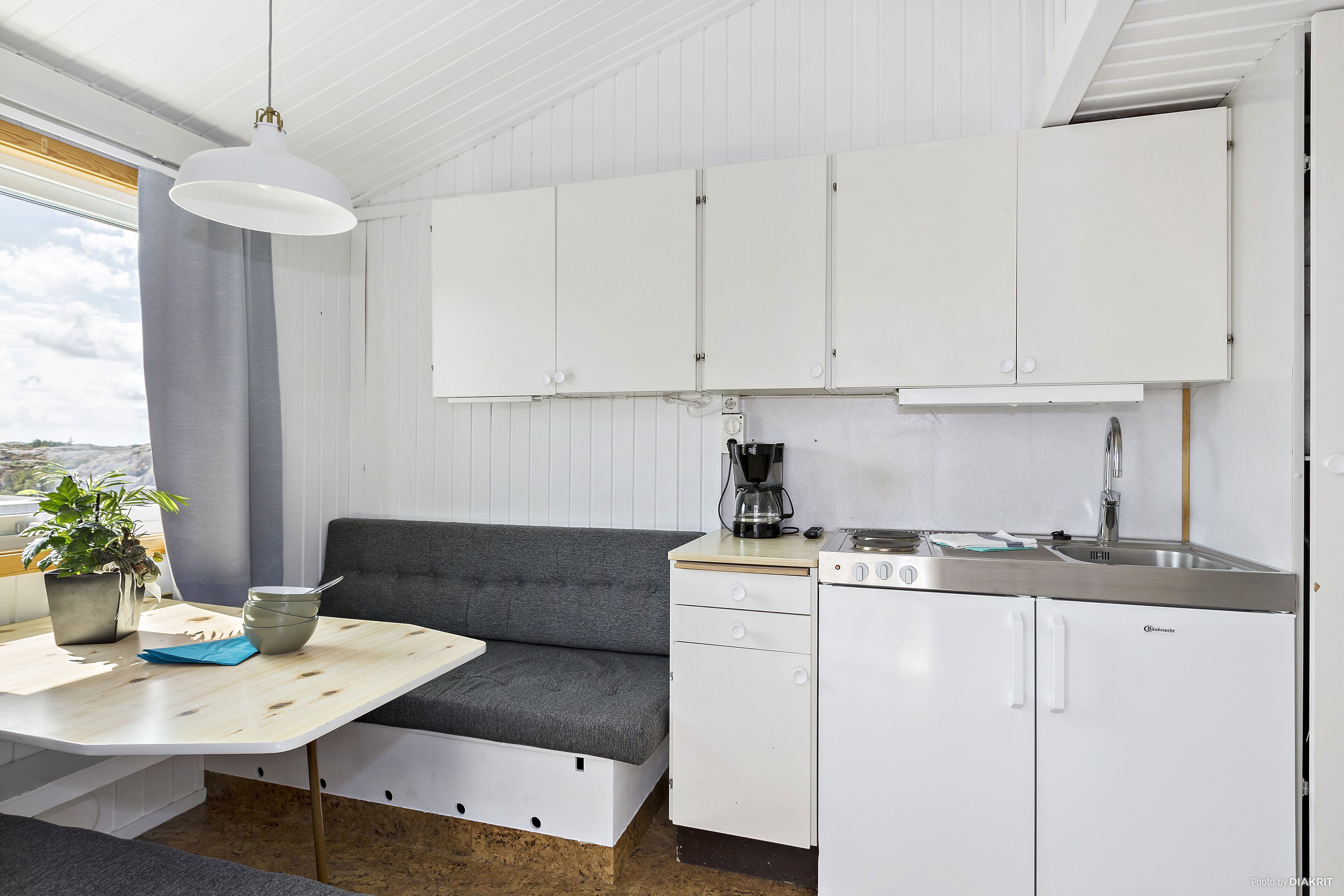 A compact kitchen with white cabinets, a grey sofa, a wooden table, and a coffee machine.