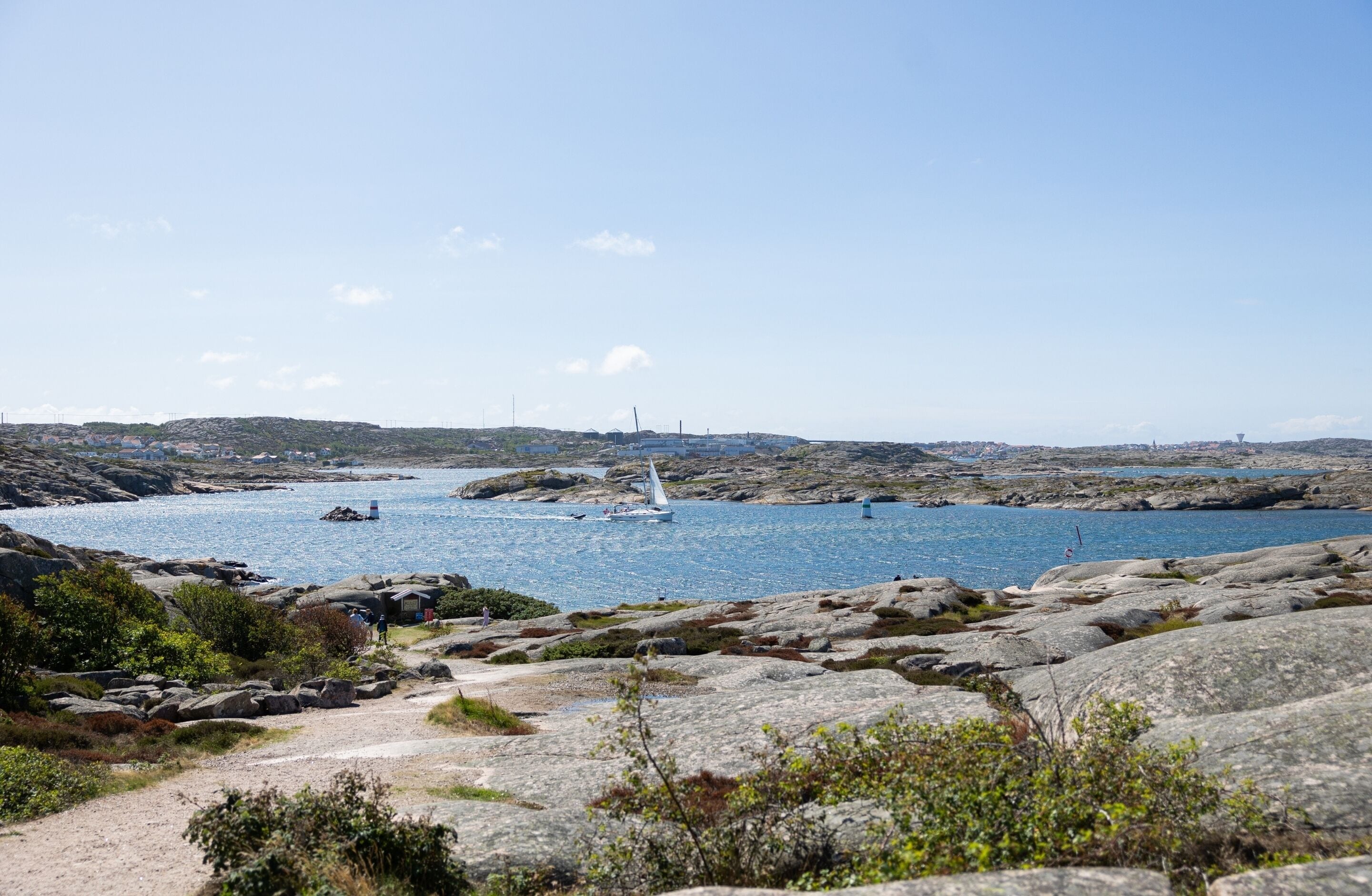A coastal landscape with rocky terrain, a clear blue sky, and a sailboat on the water.