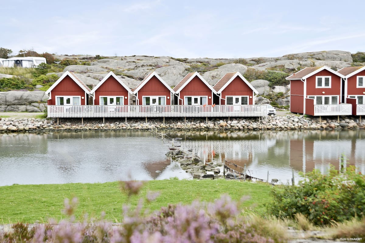 A row of red wooden cabins with white railings, situated by a calm lake with a rocky shoreline.