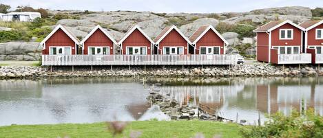 A row of red wooden cabins with white railings, situated by a calm lake with a rocky shoreline.