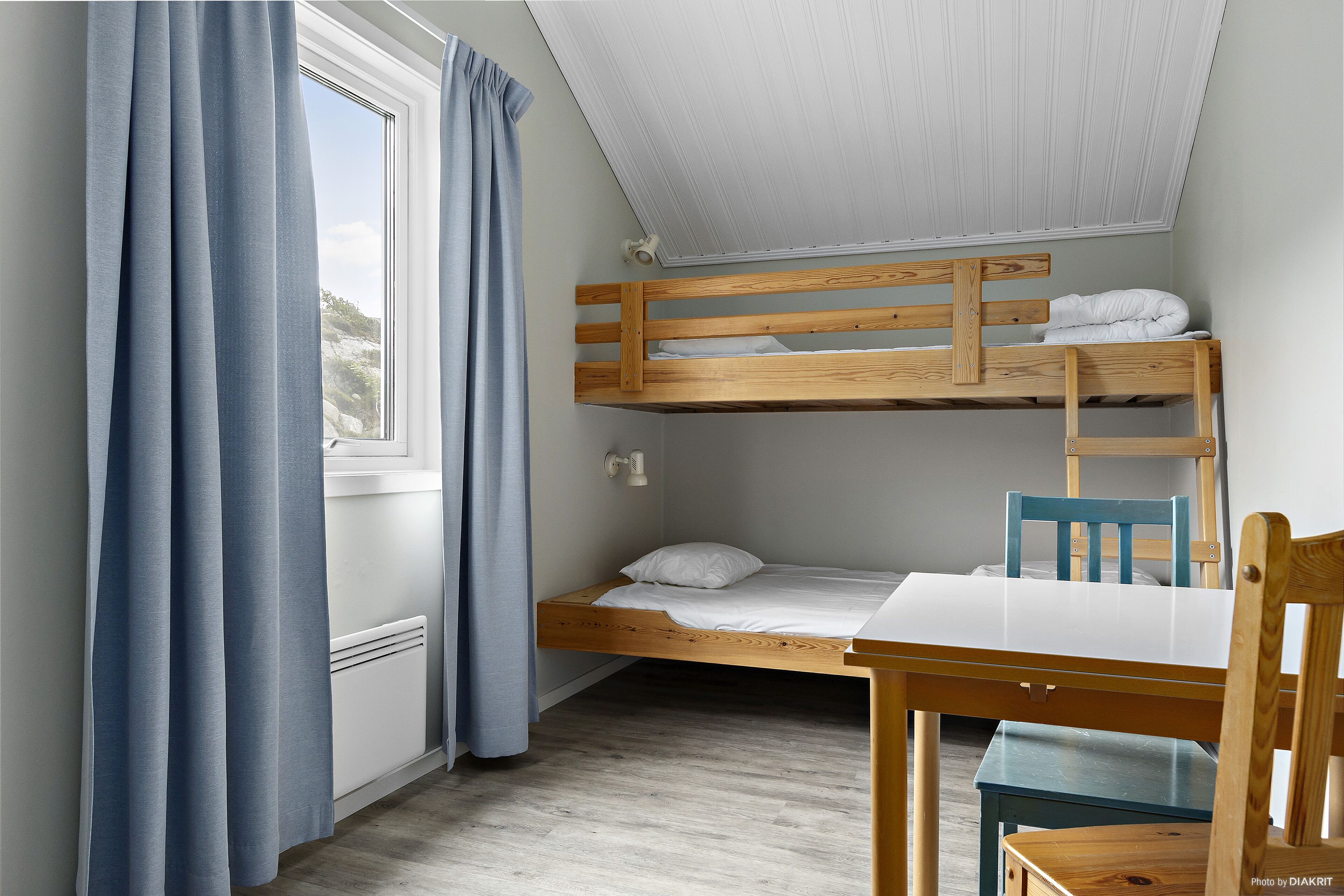 Una cama litera con estructura de madera y ropa de cama blanca, una mesita pequeña y una silla en una habitación con techo inclinado y una ventana con cortinas azules.