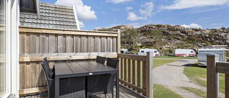 A wooden deck with a table and chairs, a wooden fence, and a view of a rocky hillside and RVs in the background.