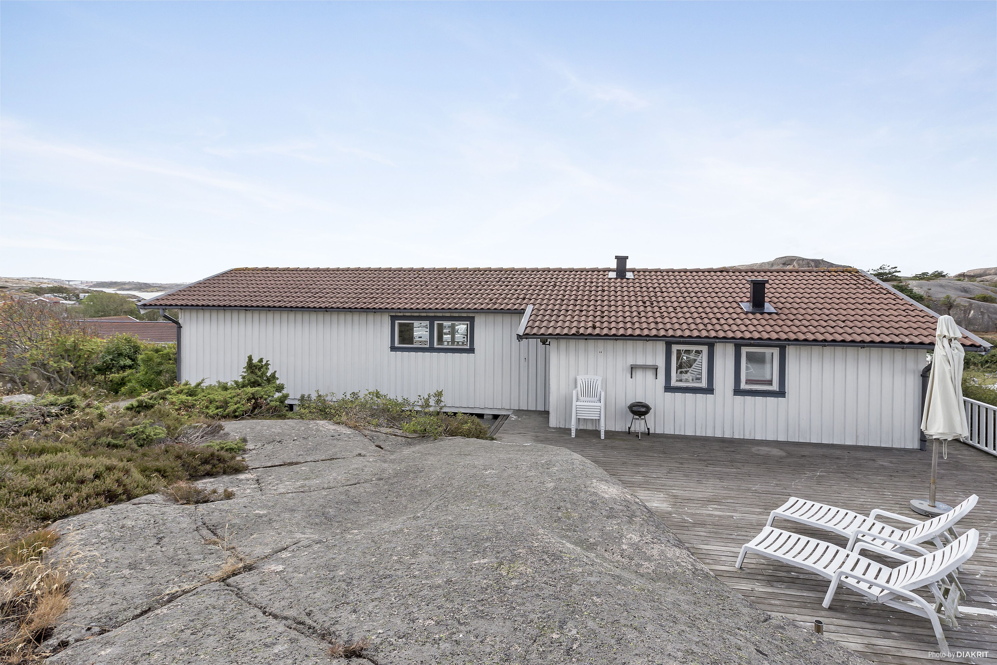 A single-story house with a tiled roof, a porch with two white chairs, and a small table.