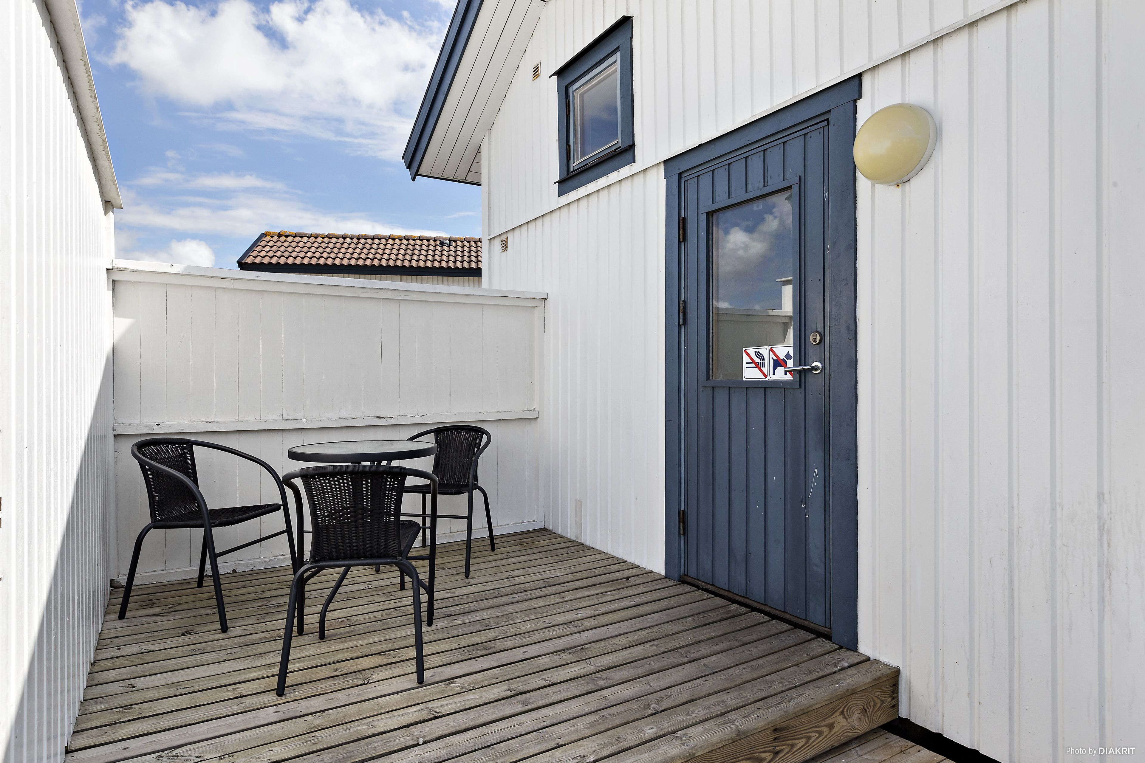 A wooden deck with a table and chairs, a blue door, and a white building with a tiled roof.