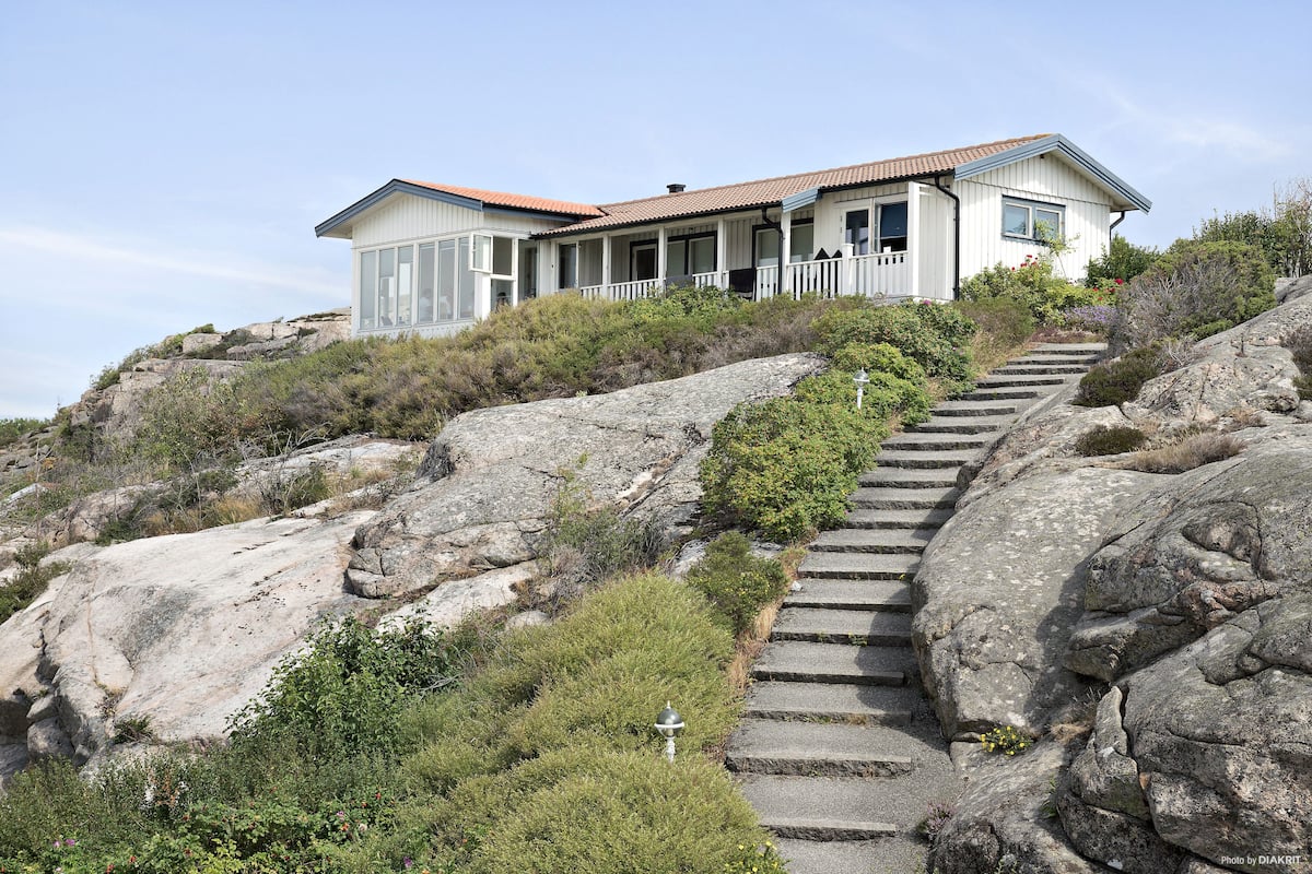 A white house with a red roof on a rocky hillside, featuring a stone staircase leading to the entrance.