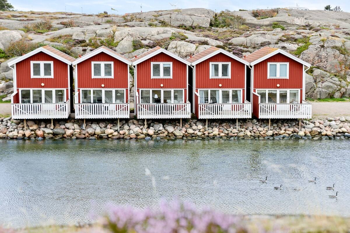 A row of red wooden houses with white railings by a rocky shore and a calm body of water.