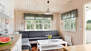 A cozy cabin-style room with a wooden interior, a kitchenette, a dining area, and large windows offering a view of greenery outside. - First Camp Kärradal (Varberg)