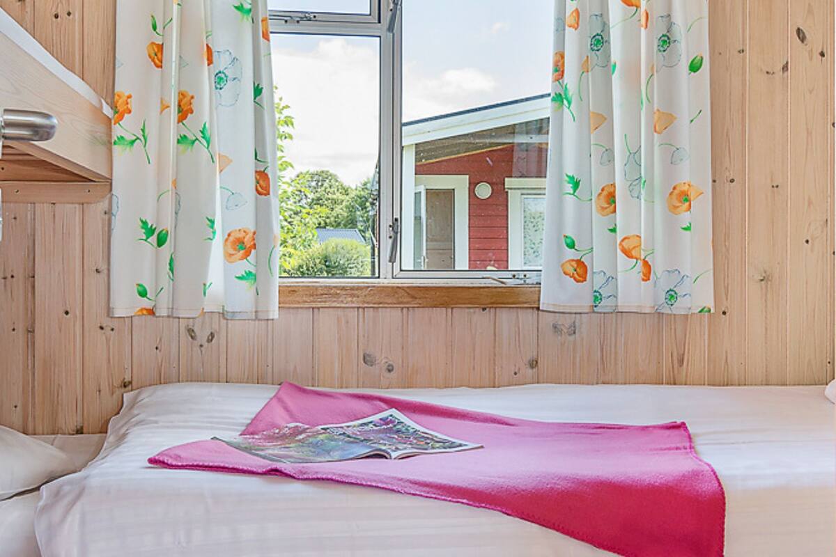 Un dormitorio con paredes de madera, una ventana con cortinas estampadas, una cama con ropa blanca y un edredón rosa.