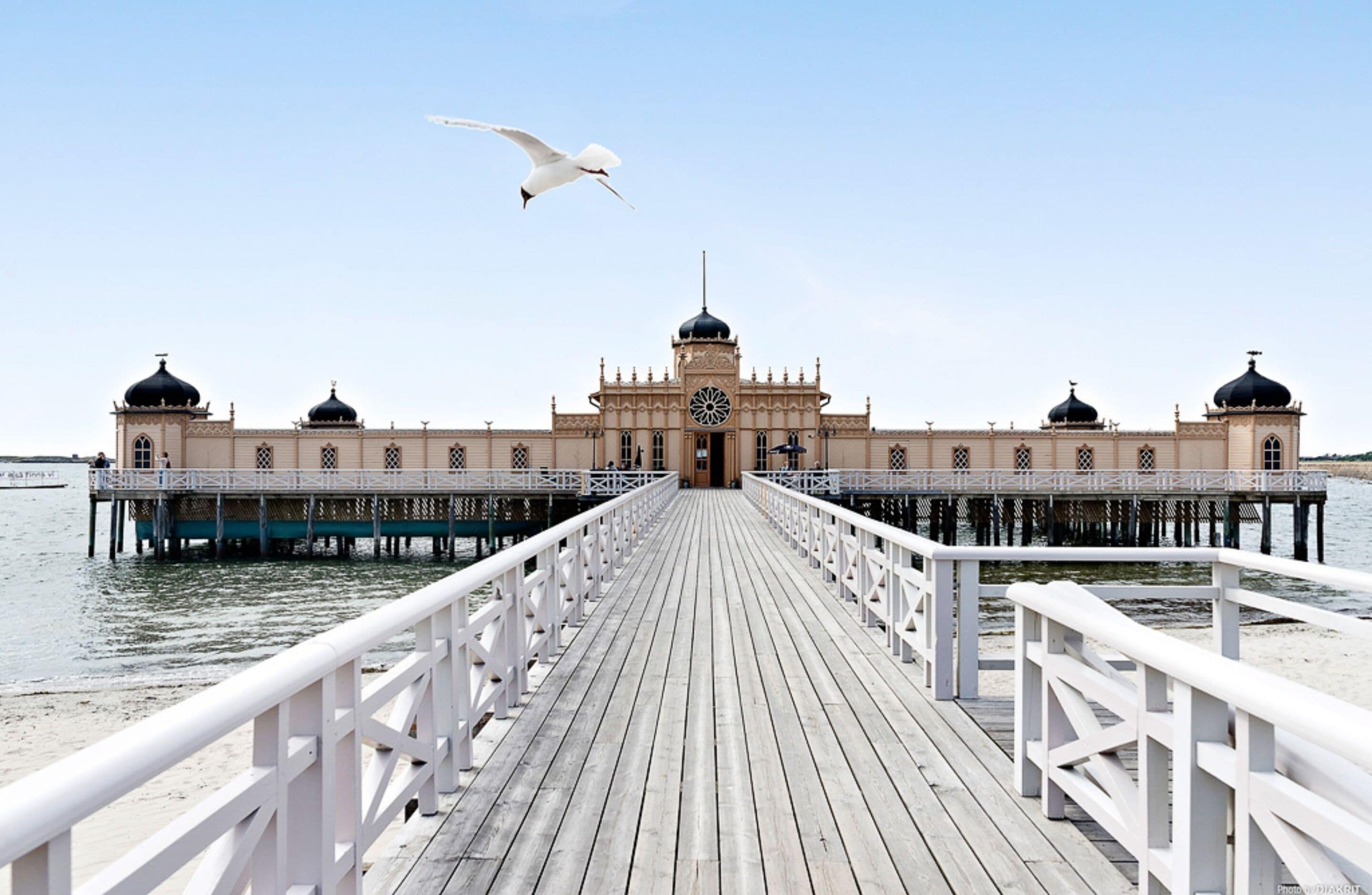A wooden pier leading to a building with domed towers and a central clock tower.