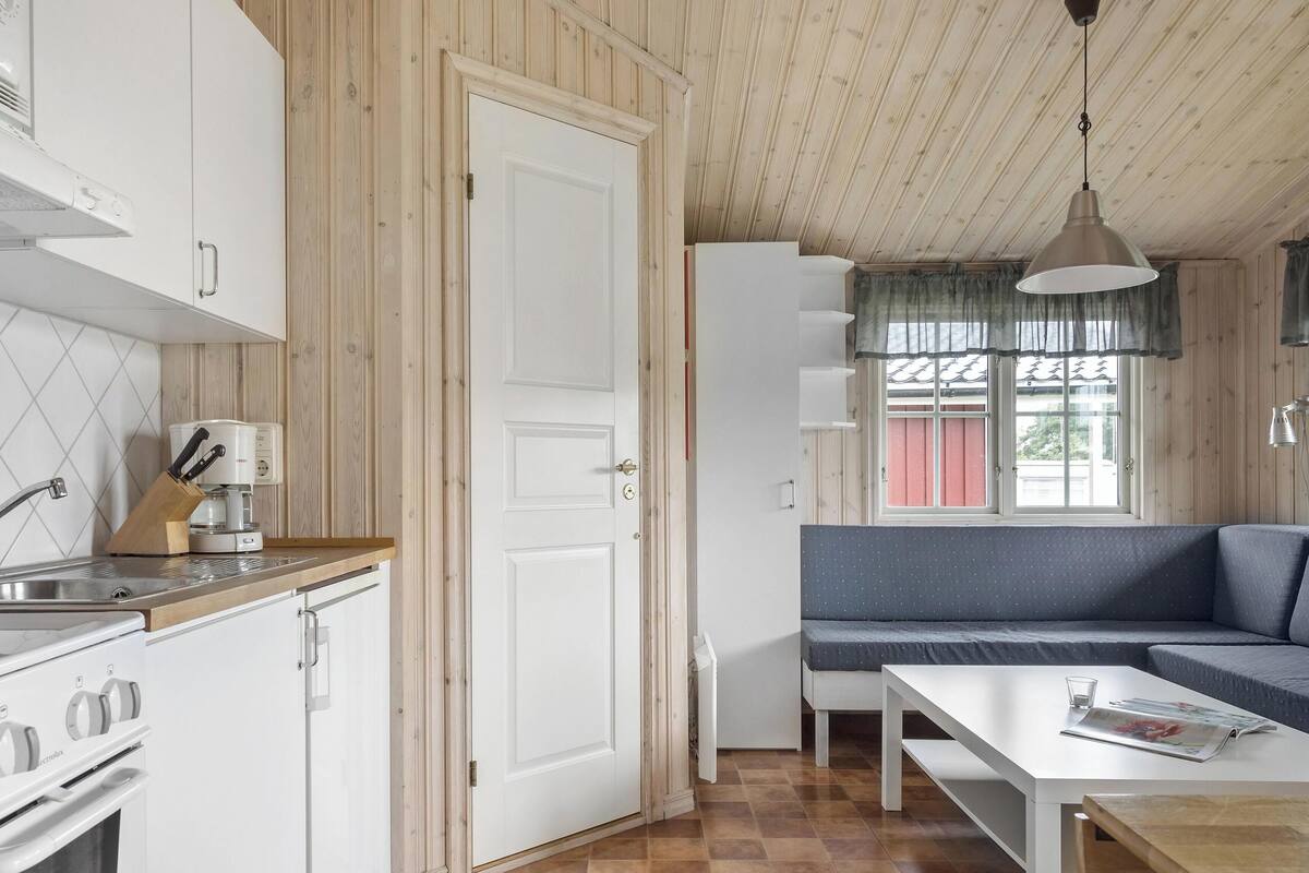 Una cocina compacta con estufa, pileta y cafetera, una puerta blanca y un sofá azul en una habitación con paredes de madera y una ventana.