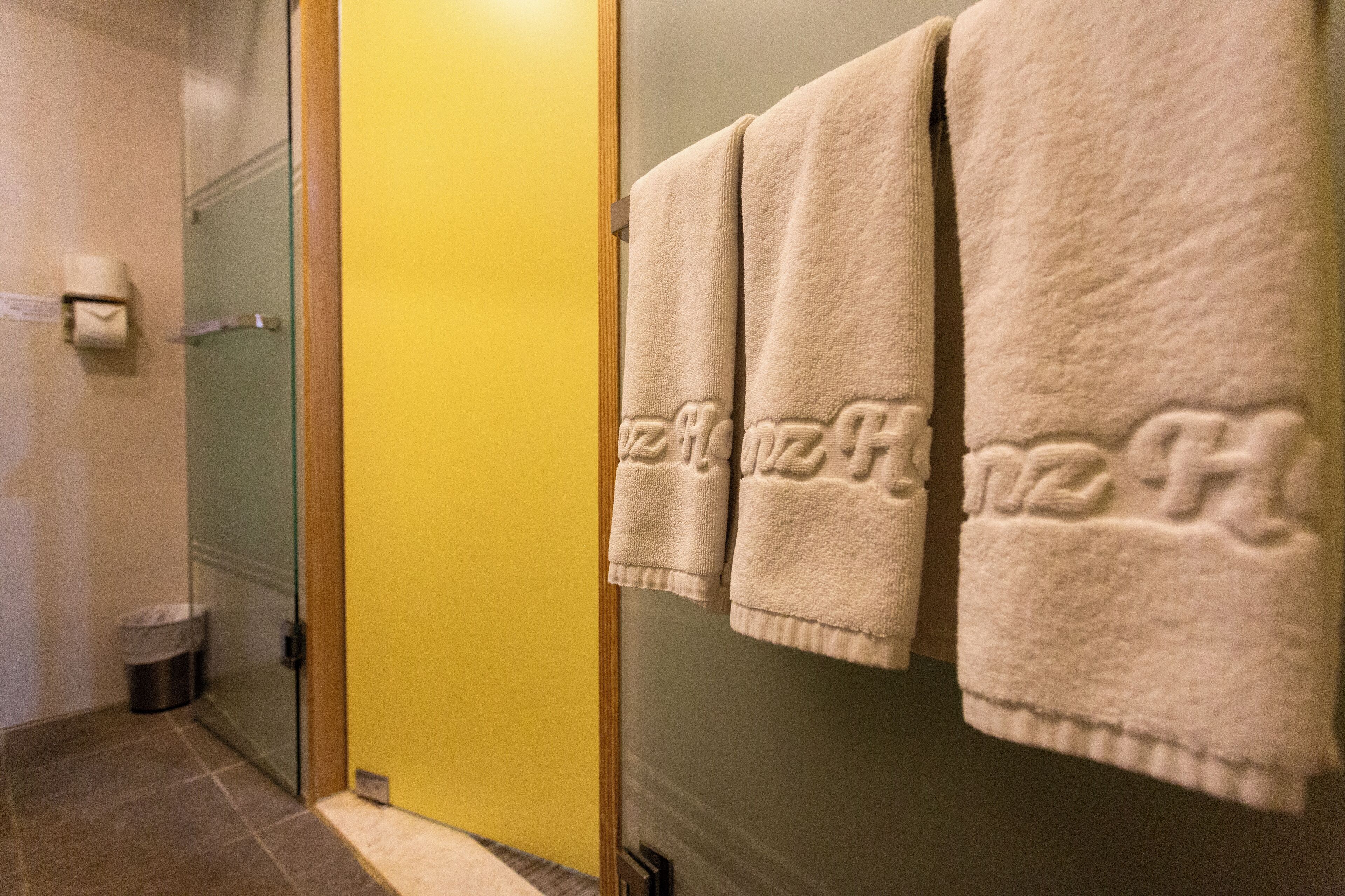 A bathroom with a yellow wall, a glass shower door, and three towels hanging on a rack.