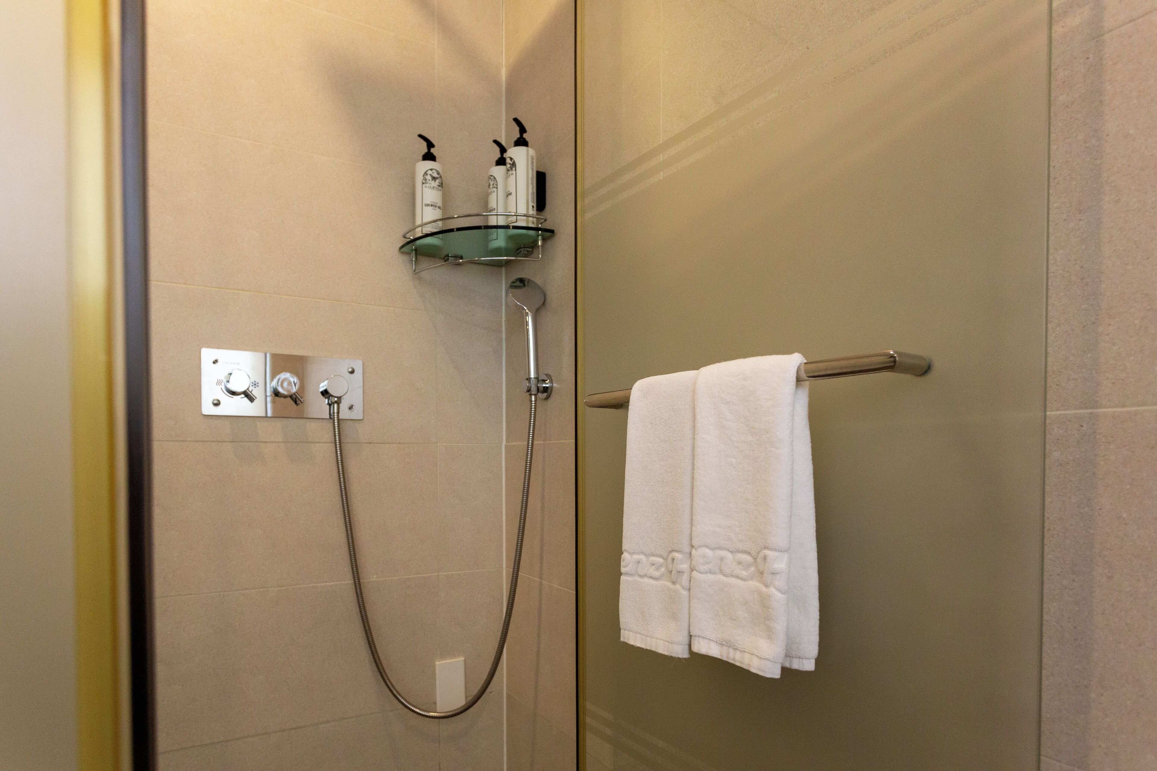A shower with a glass shelf holding toiletries, a towel rack with a towel, and a control panel.
