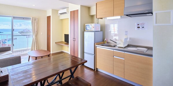 Una cocina compacta con refrigerador, microondas y pileta. Hay una mesa de madera y un pequeño espacio de descanso con vista al mar.