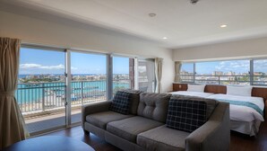 A hotel room with a large balcony, a sofa, a bed, and a view of the city and sea. - Seaside Condominium Rana Chatan (Chatan)