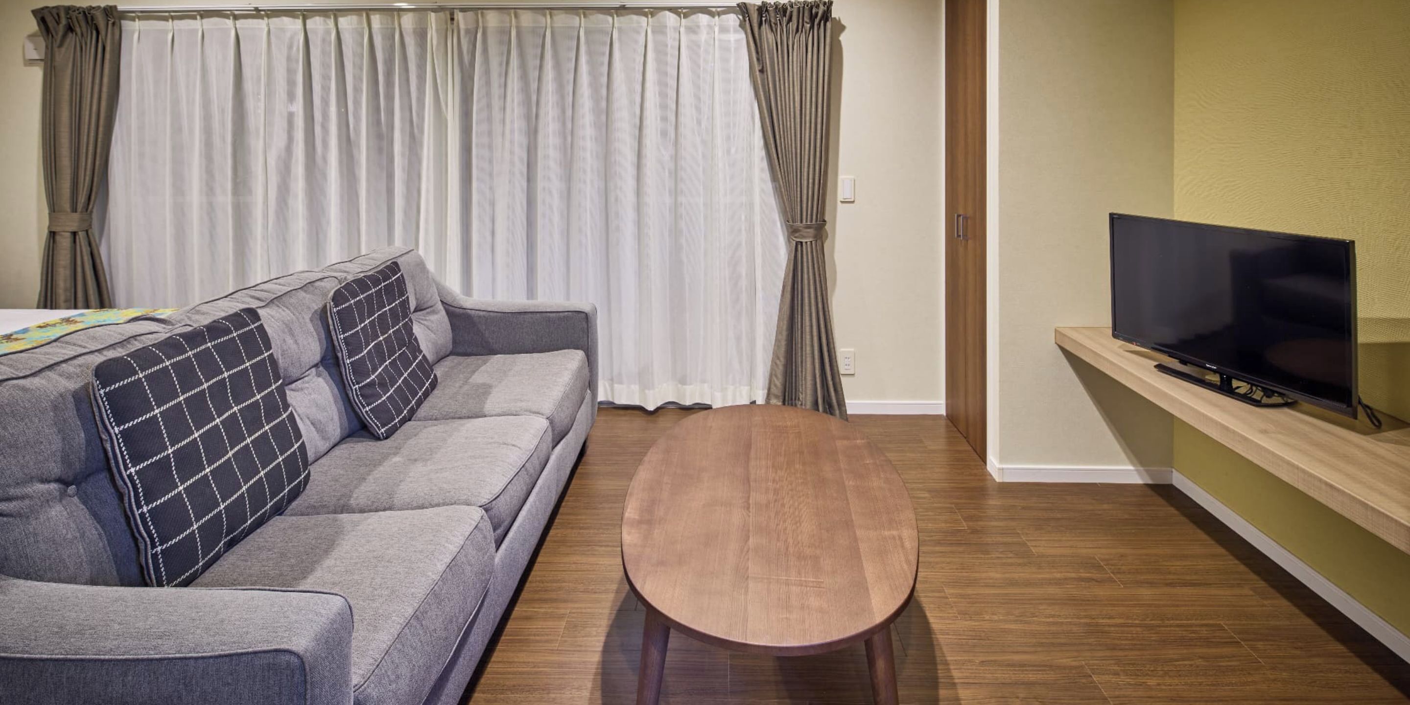 A living room with a grey sofa, a wooden coffee table, a flat-screen TV on a wooden stand, and a window with white curtains.