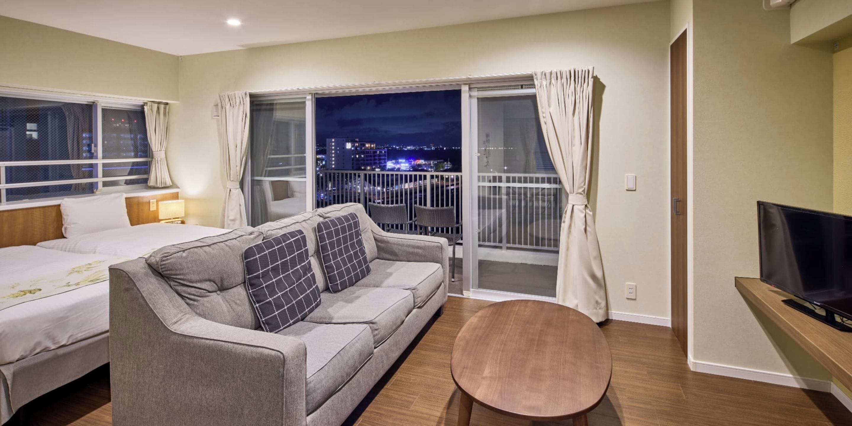 A hotel room with a bed, a sofa, a small table, a TV, and a balcony with a city view at night.