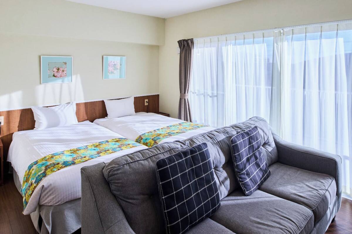 A hotel room with a bed, a sofa, a nightstand, and a window with sheer curtains.