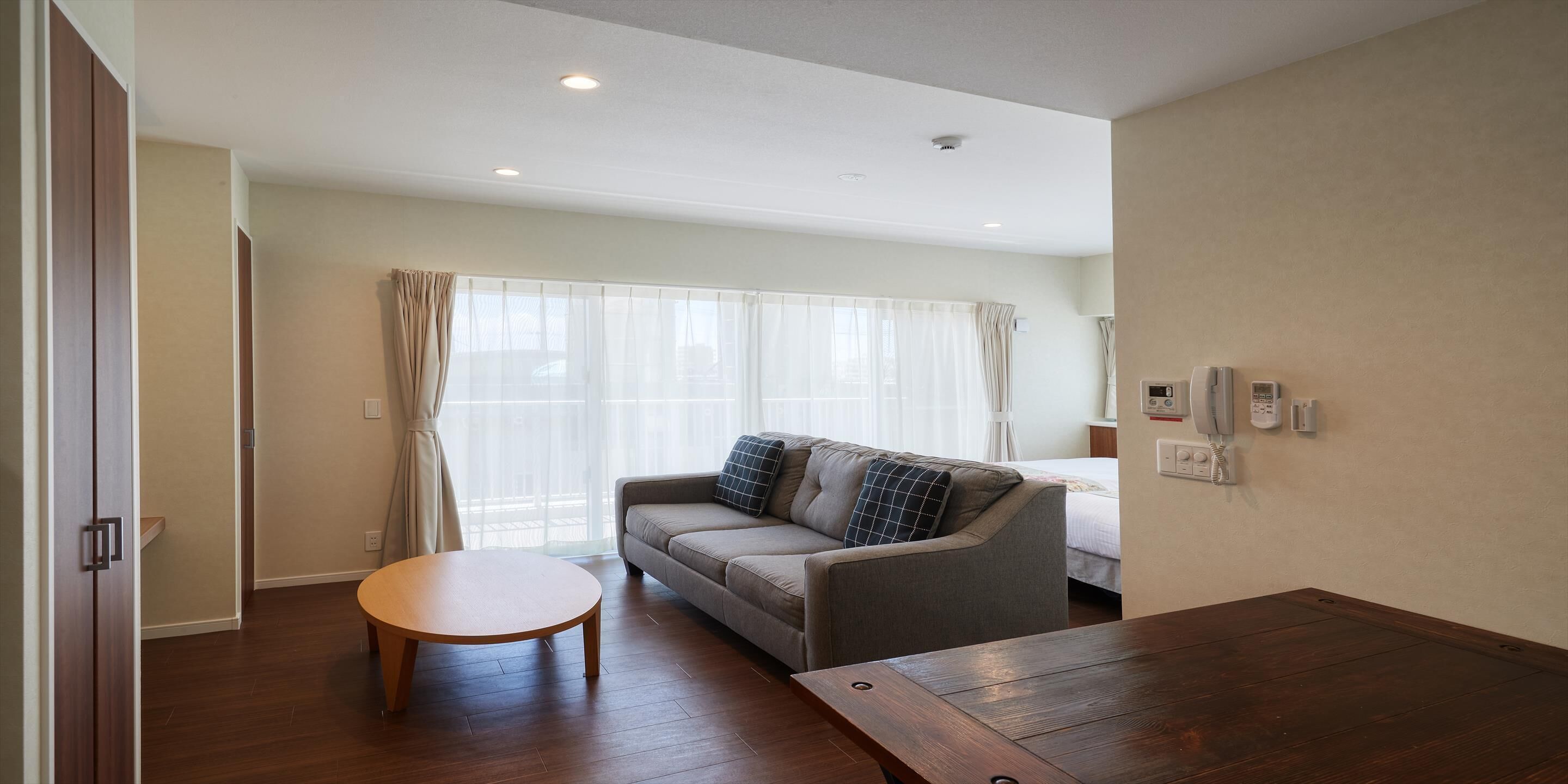 A modern hotel room with a sofa, a small round table, a wooden floor, and a large window with curtains.